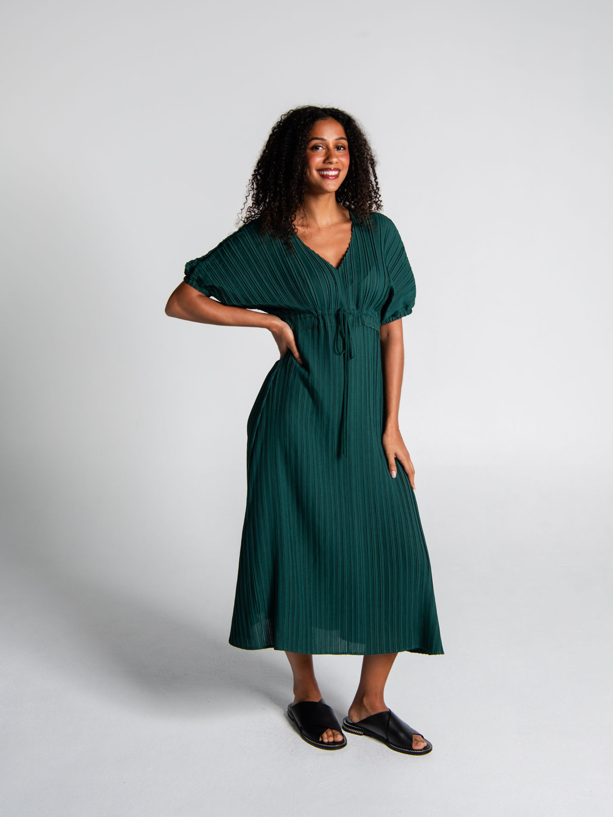 A woman modeling a dark green pleated midi dress with short puff sleeves, a V-neckline, and a matching waist tie, paired with black slide sandals against a light gray studio background.