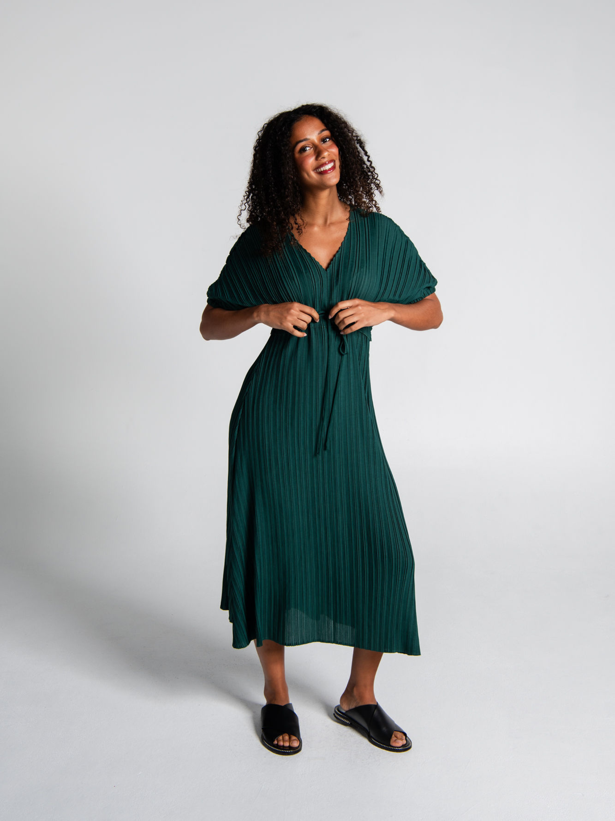 A woman modeling a dark green pleated midi dress with short sleeves and a tie-front waist, paired with black slide sandals, against a light gray studio background.